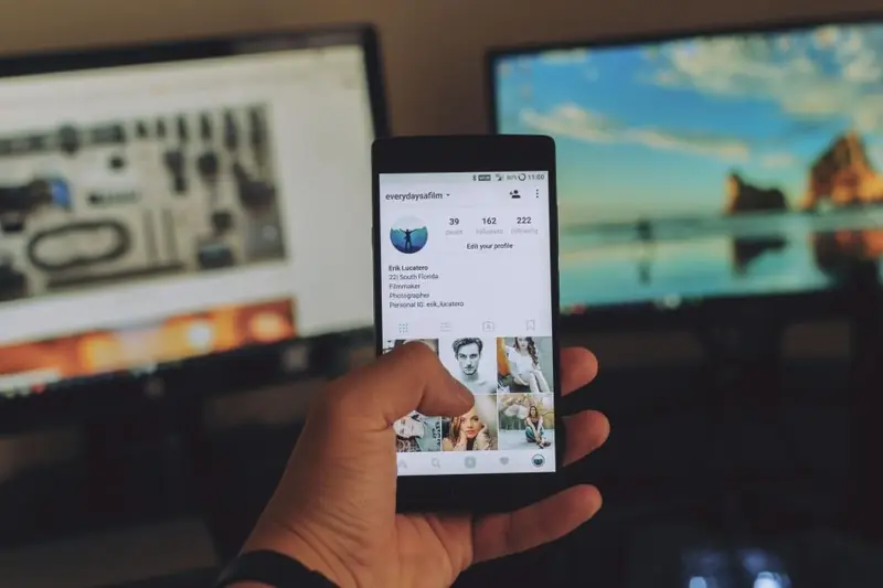 Mão segurando um celular com um perfil de rede social aberto diante de dois monitores