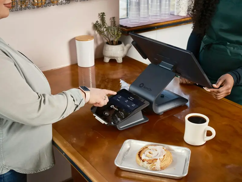 Cliente pagando com o celular no balcão de um pequeno salão de beleza, com a profissional recebendo o pagamento