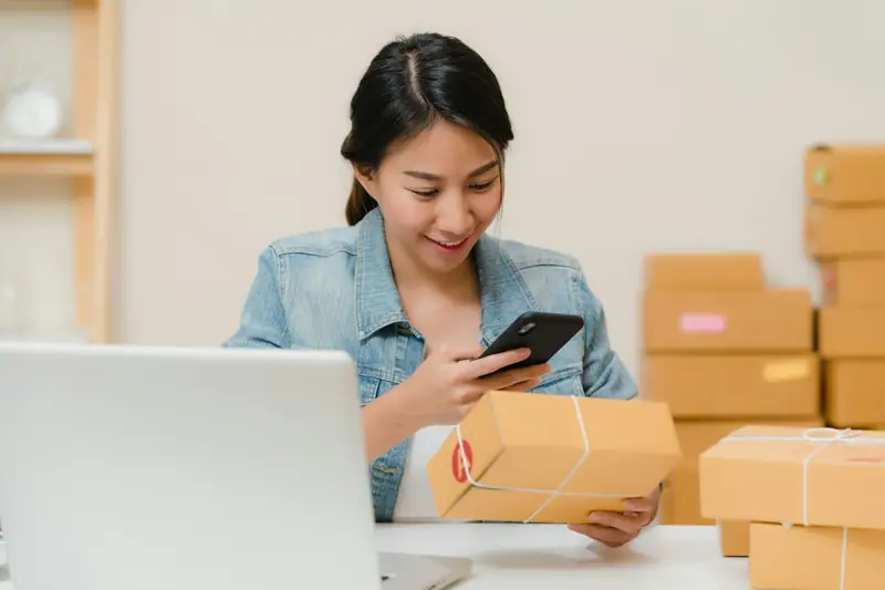 Pessoa embalando pedidos de loja online em uma mesa com caixas, etiquetas e fita adesiva